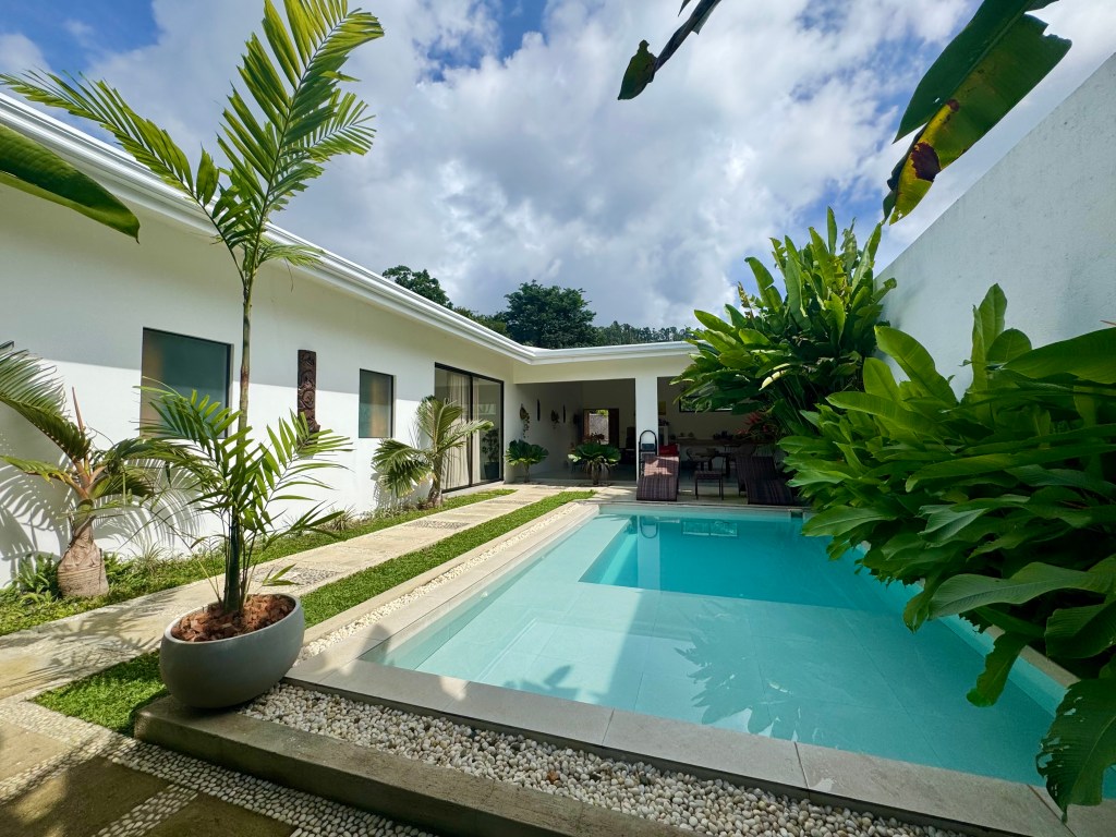 A modern outdoor area featuring a swimming pool surrounded by tropical plants and a white wall, with a covered seating area in the background under a partly cloudy sky.