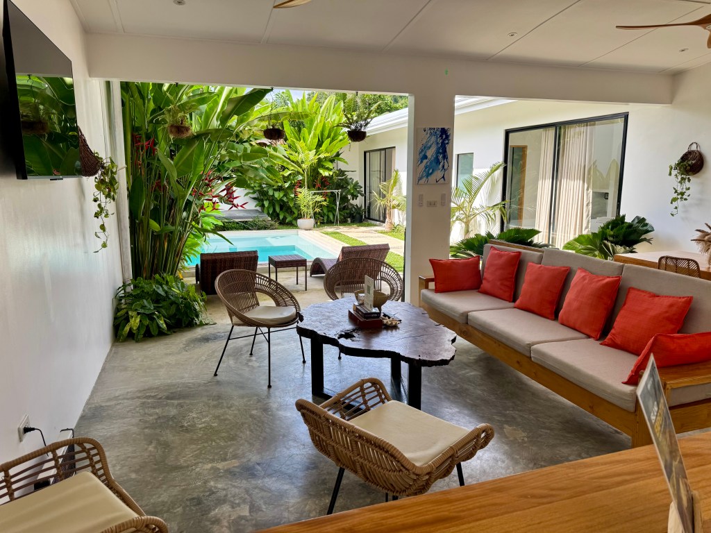 A modern living area with a view of a pool surrounded by tropical plants. The space features a comfortable sectional sofa with red cushions, a wooden table, and rattan chairs. A flat-screen TV is mounted on the wall, enhancing the inviting atmosphere.