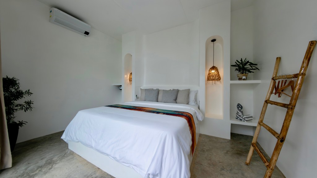 A modern bedroom featuring a white bed with decorative pillows, a colorful throw, and natural decor elements including a plant and a wooden ladder against a white wall.